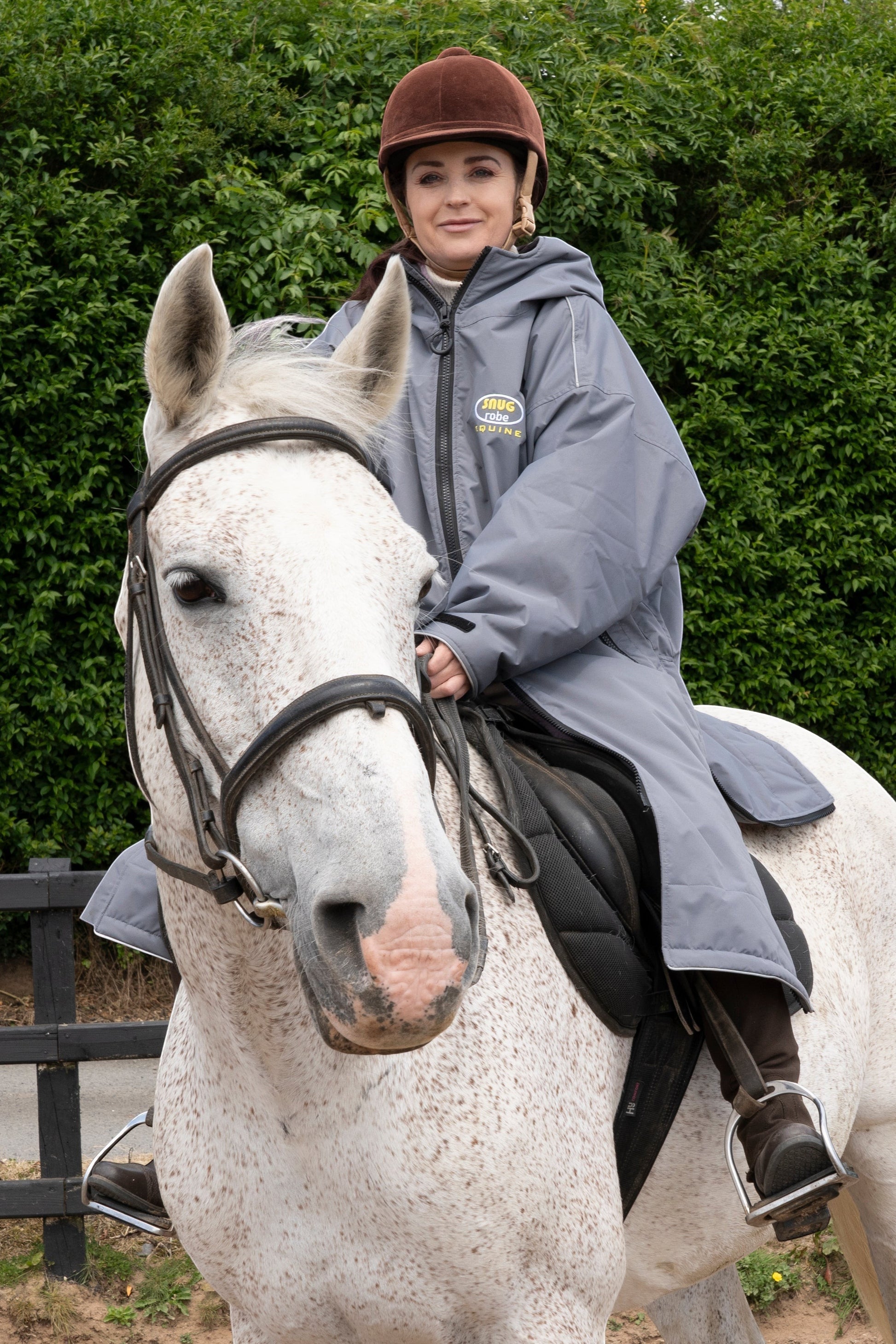 Snugrobes Equine grey waterproof robe with fleece lining worn by a woman on a horse. Features include a hood, zipped pockets, and side zips for riding.