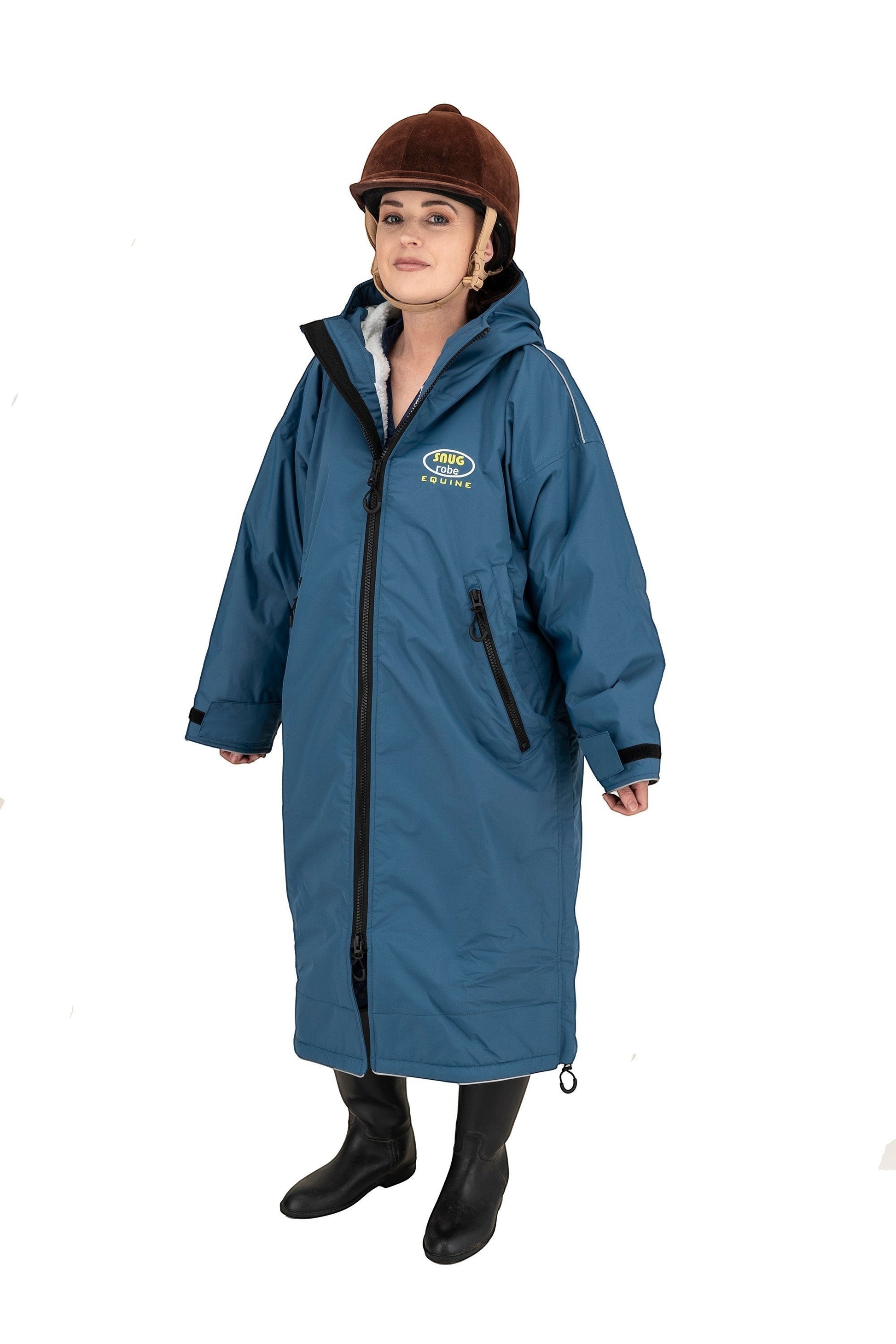 Snugrobes Equine waterproof robe in teal blue with fleece lining, hood, and reflective piping. Model wears riding helmet and boots.